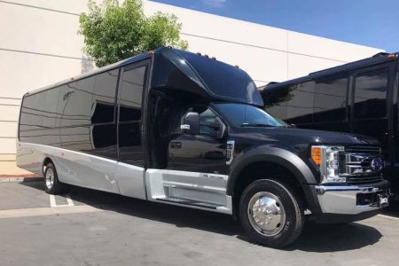 A large black and silver bus-style limousine is parked in a lot next to a beige building on a sunny day.