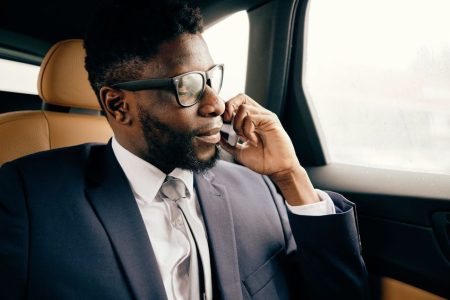 Man in a suit and glasses sits in a car’s back seat, talking on a smartphone and looking out the window.