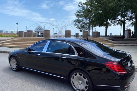 A black luxury sedan is parked near a stone fountain and steps under a partly cloudy sky.