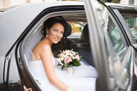 Bride in white dress holding bouquet, sitting in the back seat of a car with the door open, looking at the camera.