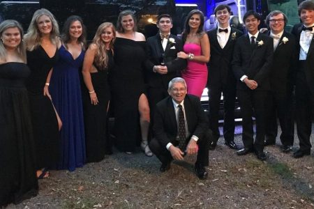 A group of young adults in formal attire poses outdoors in front of a black vehicle, with one older man kneeling in front.